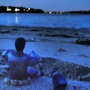 Armored space soldier on the beach at night with helmet beside him facing the waves