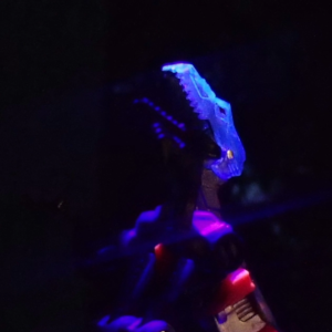 A robotic velociraptor looks upwards as its armor plating is lit up by blue light.