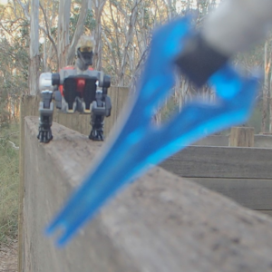 An action shot of a white-armored space arm holding an energy sword with a robotic velociraptor approaching menacingly, posed on a wood fence in a forest.