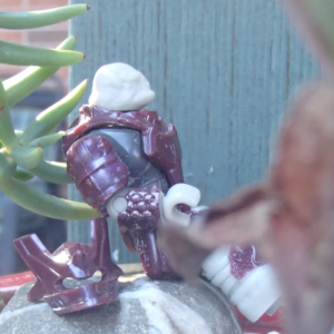 A crimson-armored space alien sits on a stone next to its helmet amidst white pebbles and succulents.