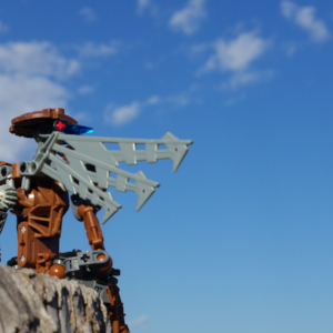 A brown Bionicle with robotic wings perched on a wooden fencepost looking over a field on a clear day.