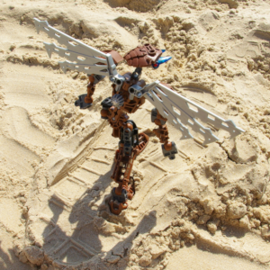 High angle view of a brown Bionicle with robotic wings walking on the beach in the middle of a human flip-flop footprint.