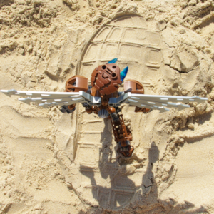 Top-down view of a brown Bionicle with robotic wings walking on the beach in the middle of a human flip-flop footprint.