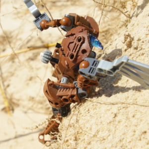 Top-down view of a brown Bionicle holding robotic wings to climb up a steep sand cliff.