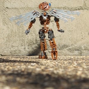 A brown Bionicle with robotic wings steps towards a brick wall while looking far upwards.