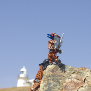 Low angle shot of a brown Bionicle with robotic wings perched on a rock with a lighthouse on a green hill in the background.